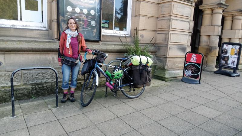 A woman in blue jeans, red jacket and pink sweater is standing holding the handlebar of a packed touring bike that is leaning against a cycle hoop. They are outside a big stone building