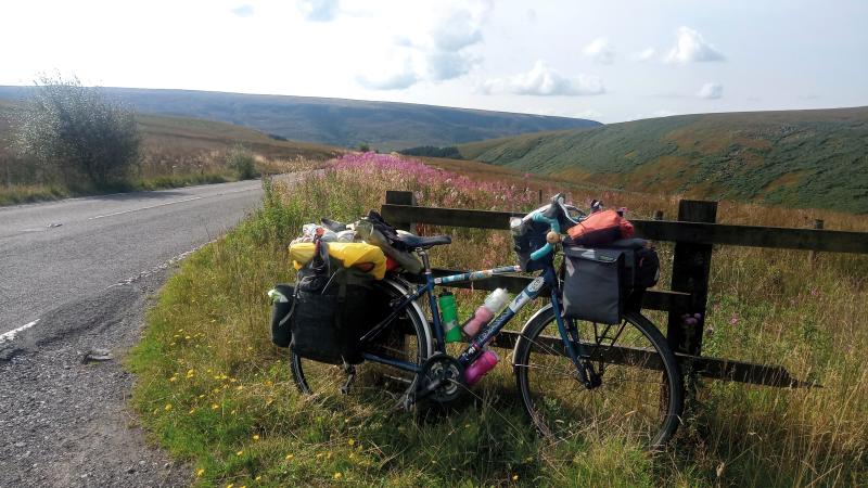 A loaded touring bike is leaning against a wooden fence next to a country road. In the background is the high moorland of Holme Moss