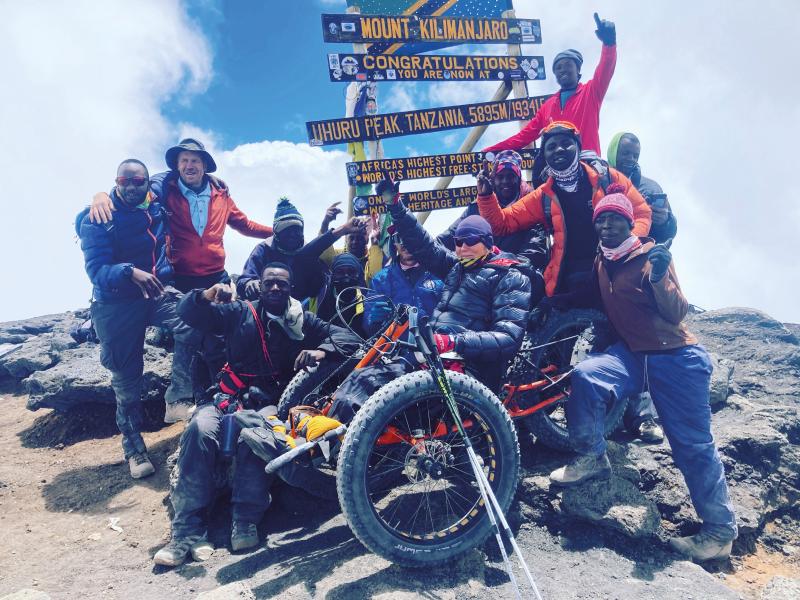 A mixed group of people are standing around a sign saying they've made it to the top of a mountain. They're all celebrating. A woman on a fat trike is in the centre