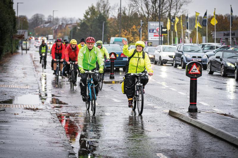 A group of people are cycling along a very rainy protected cycle path. They are wearing waterproofs, but are smiling and seem to be in good spirits. In the background is a stationary line of traffic