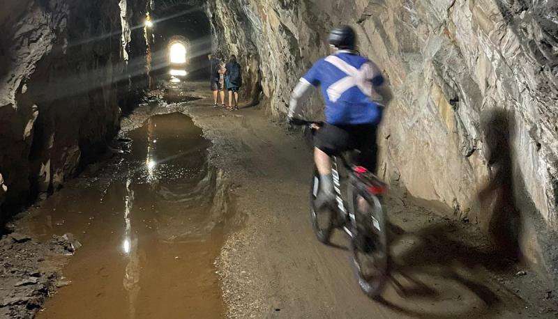 A man in a blue and white cross of St Andrew jersey, black shorts and cycling helmet is riding a touring bike through a tunnel with a stream alongside