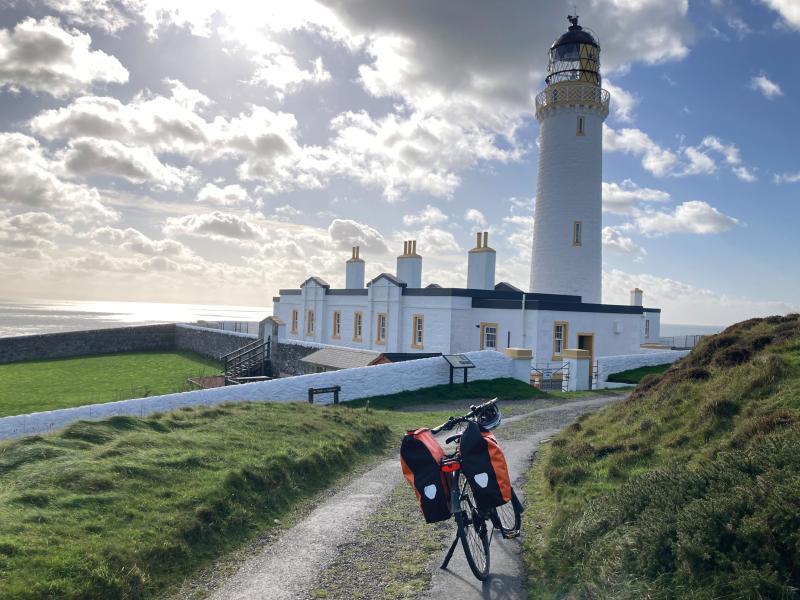 A packed touring bike with two big panniers is propped on its kick stand on a track facing a white lighthouse and building
