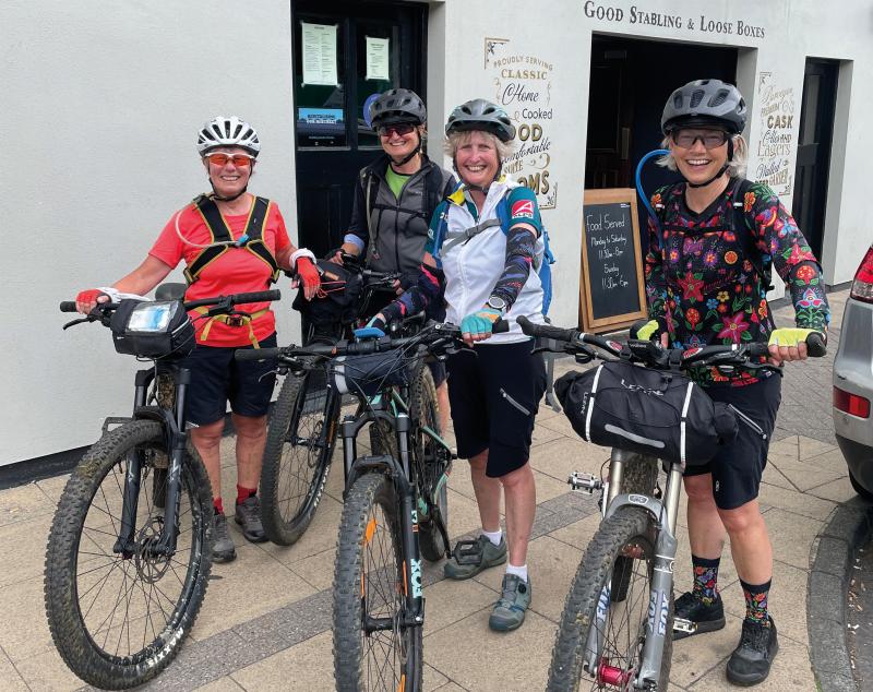 Four women with loaded mountain bikes are standing outside a pub. They are wearing MTB clothing and cycle helmets. They are smiling at the camera.