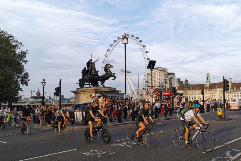 People are cycling across a road on the south bank of the Thames. Pedestrians are waiting to cross the road. The London Eye is in the background