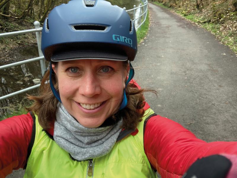 Victoria Lloyd-Gent has taken a selfie. She is wearing a blue cycle helmet, a red cycling jersey and yellow gilet and a grey buff. She has long hair and is smiling at the camera