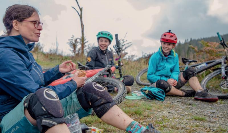 Three people are sitting on the ground. It's muddy and they're wearing muddy mountain biking kit. Their bikes are lying on the ground next to them. One woman is holding a pie.