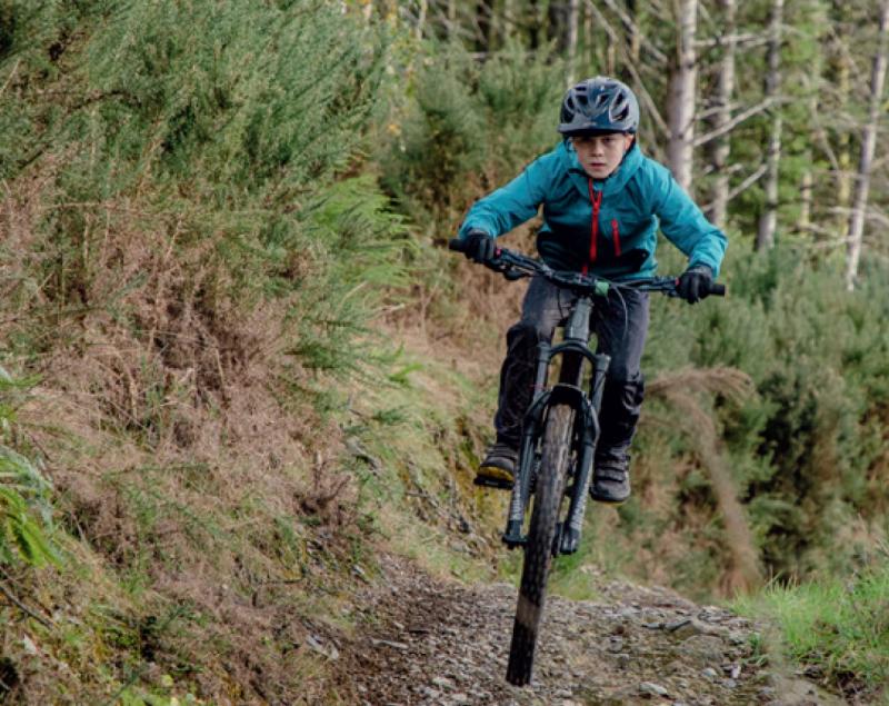 A boy is cycling on a mountain bike down a gravel trail. He is wearing a blue waterproof cycling jacket and black mountain biking trousers and a helmet.