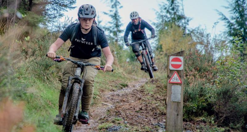 Two people are riding down a very muddy off-road track on mountain bikes. They are wearing mountain biking kit and helmets.