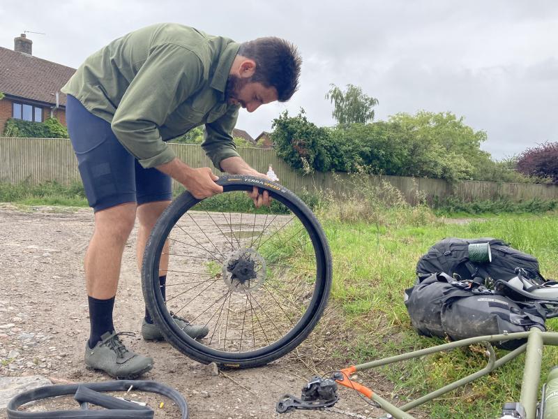 A man is fitting a bike tyre to his rear wheel, an inner tube is on the ground beside him and you can just see the frame of his bike leaning on the ground