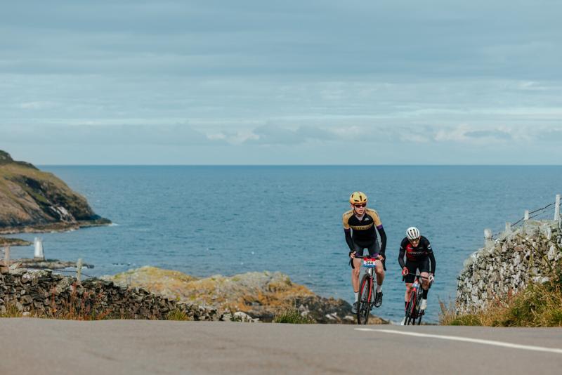 Two men are cycling up a coastal road with the sea and cliffs in the background. They are wearing cycling kit