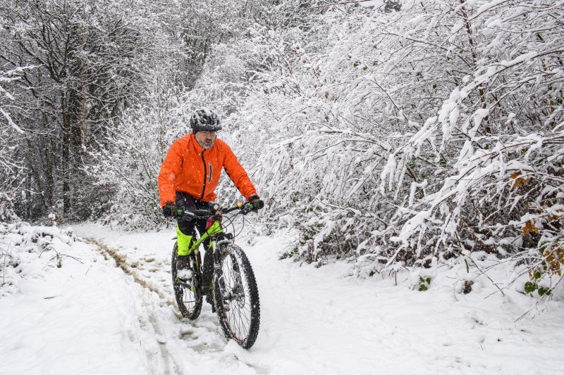 A man on a mountain bike is riding through a snowy off-road scene. He is wearing a bright orange winter jacket and black cycling trousers