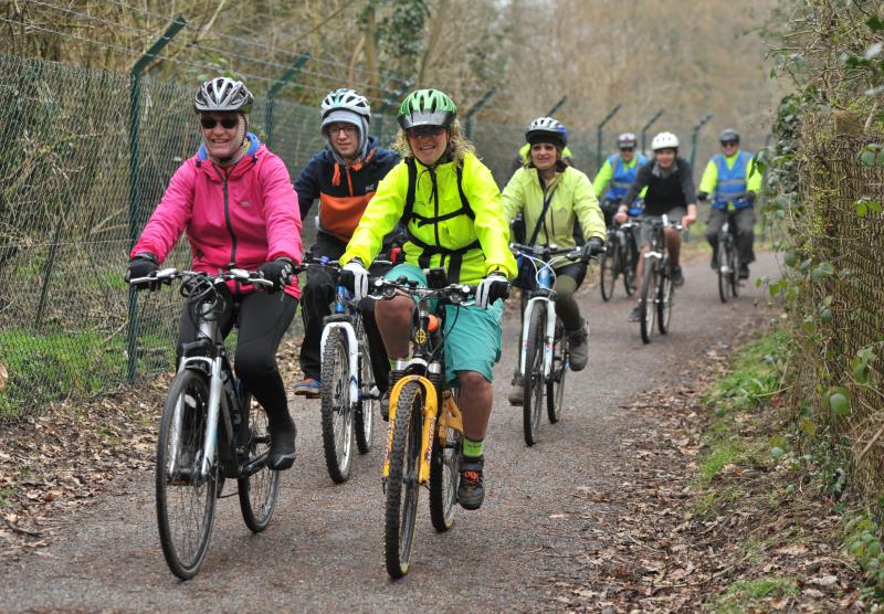 A group of people are cycling on an off-road track. They are mostly on mountain bikes. They are wearing a mix of hi-vis and normal clothing. They are smiling