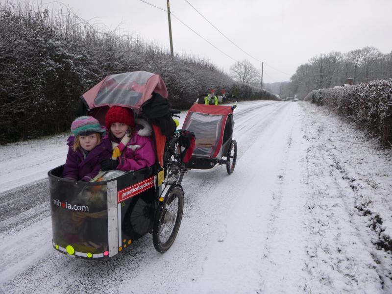 A cargo bike with a trailer on the back and child box on the front with two young children in. It's a very wintry scene with snow on the road and in the hedges and trees. The children are wrapped up warm.