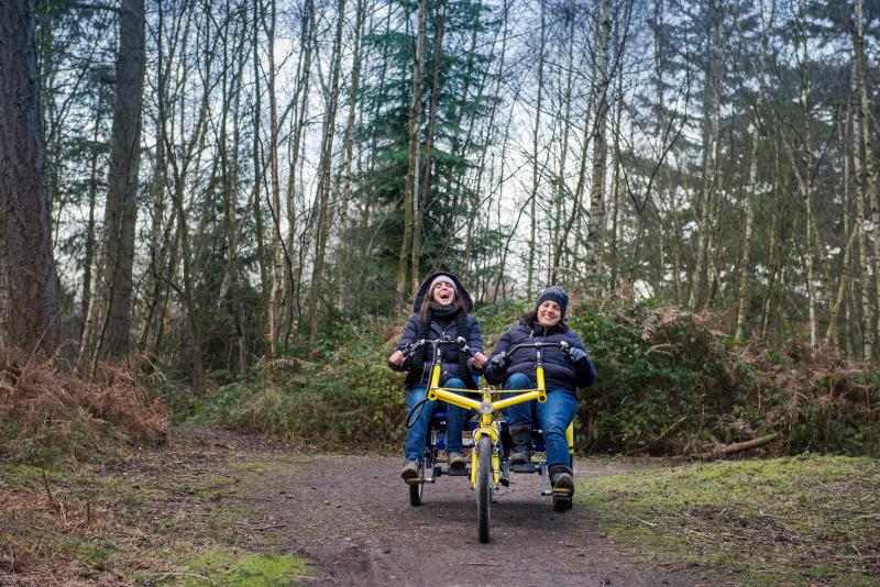 Two women cycling on a two seater trike outside in the woods on a slightly muddy footpath
