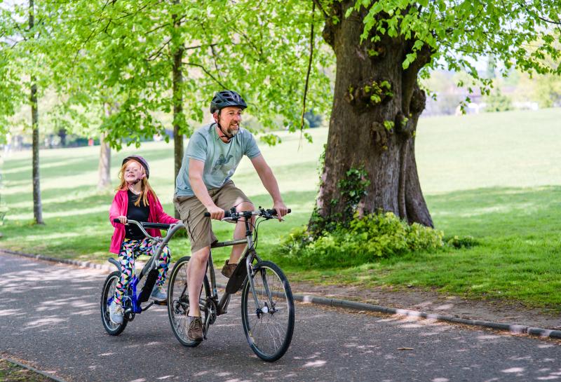 A man is cycling along a tarmac path in a park. He is wearing shorts and T-shirt. There is a tag-along bike attached to his bike and a young girl is cycling on it.