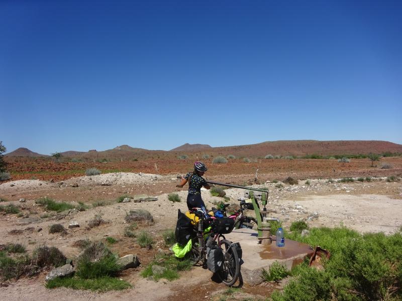 A woman in cycling kit is operating a well in Africa to fill a large water bottle. A loaded touring bike is propped against the well's concrete plinth