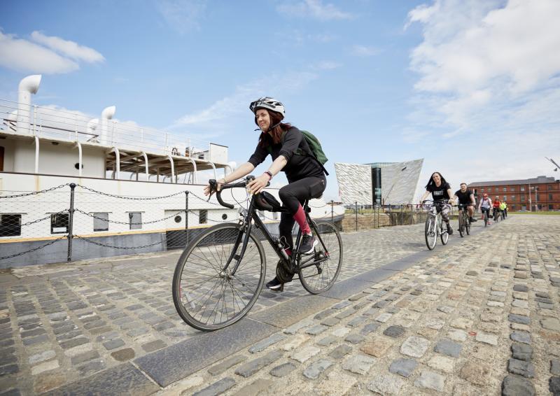 A woman riding on a bike is leading a group of riders along a stone path at some docks. On the rider’s right is a boat and behind them a red brick building