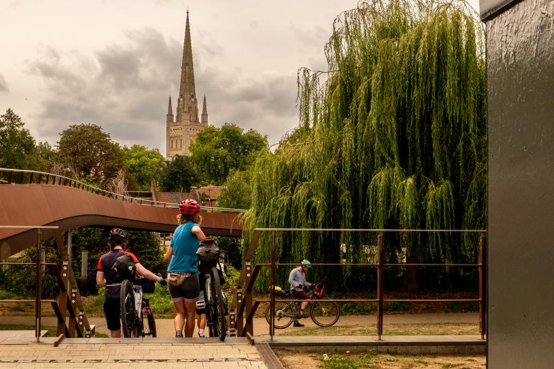Cyclists are wheeling their bikes down some steps. In the background is Norwich Cathedral.