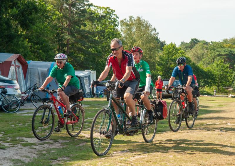 A group of people are cycling through a field with tents lined up along the left-hand side. Two, a man and boy, are on a green tandem. There is also a woman on a mountain bike and a man on a road bike.