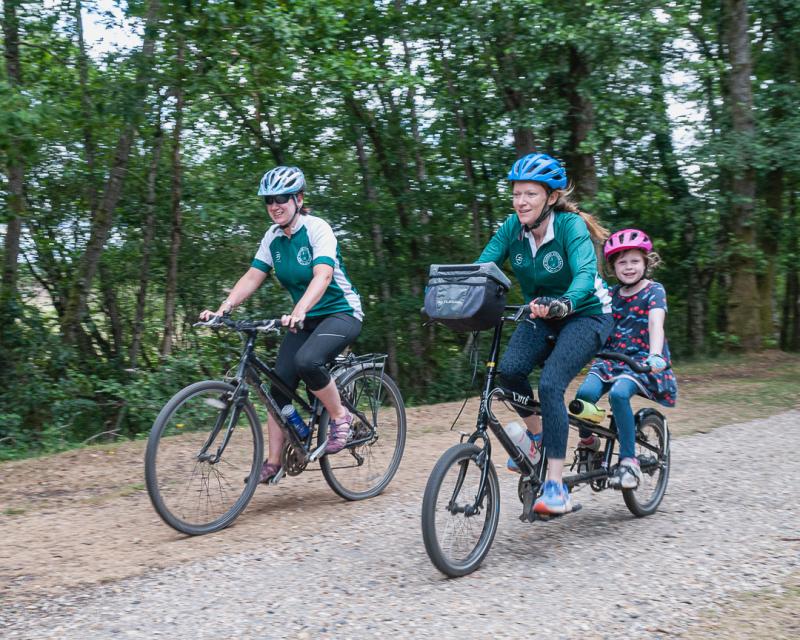 A woman and a child are on a black tandem with small wheels riding on an off-road gravel track. They are cycling next to a woman on a black hybrid bike. The two women are wearing club kit. They all have helmets