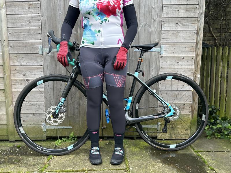 A woman in cycling jersey and shorts with Van Rysel arm and leg warmers is standing in front of a Felt road bike leaning against a garden shed