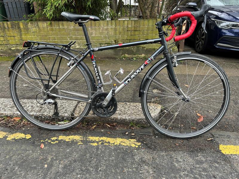 A black Dawes Galaxy touring bike with red bar tape is propped against the curb with the front wheel in a pothole