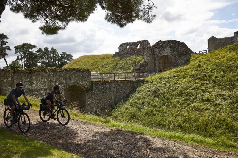 Two people on packed touring bikes are cycling past the ruins of a castle