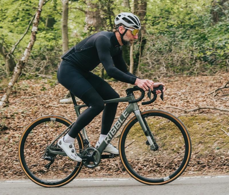 A man is riding an olive green e-road bike along a country road in a wood. He is wearing full cycling kit and a helmet.