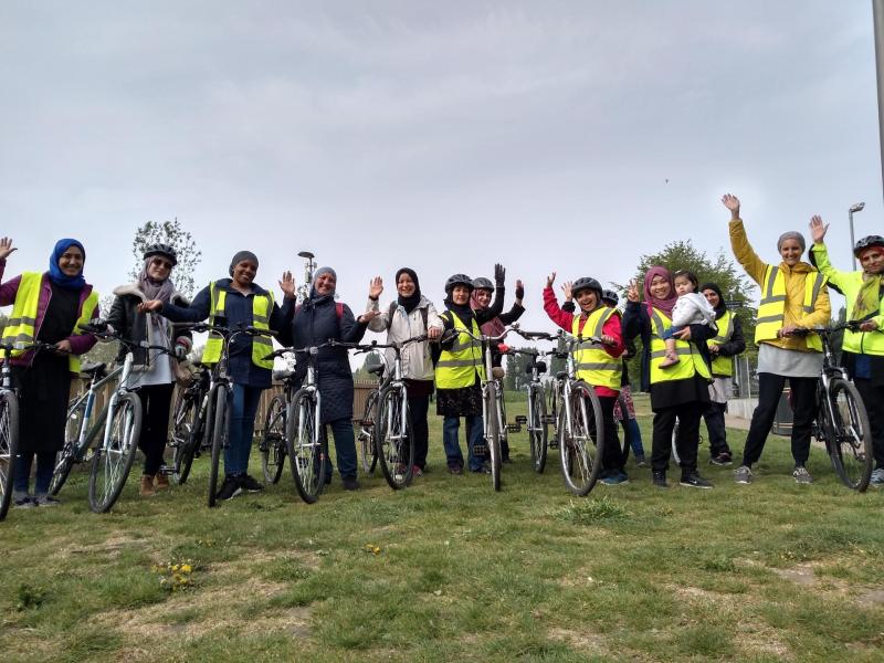 A big group of women is standing on a field all holding bikes. They are all wearing headscarves and are smiling. One is holding a baby. They are waving at the camera