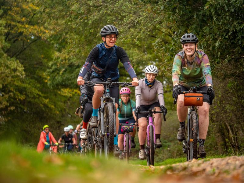 A group of women are cycling in a muddy forest. They're wearing mountain biking kit and on a mix of gravel and mountain bikes