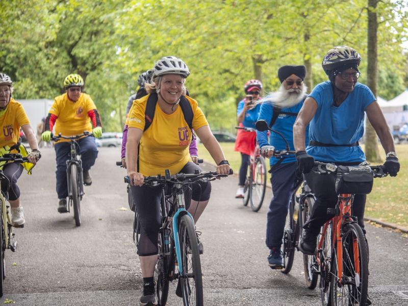 A group of people is riding along a paved, tree-lined path. They are mostly on hybrid or mountain bikes. Some are wearing yellow T-shirts with a Pumped Up Crew logo.