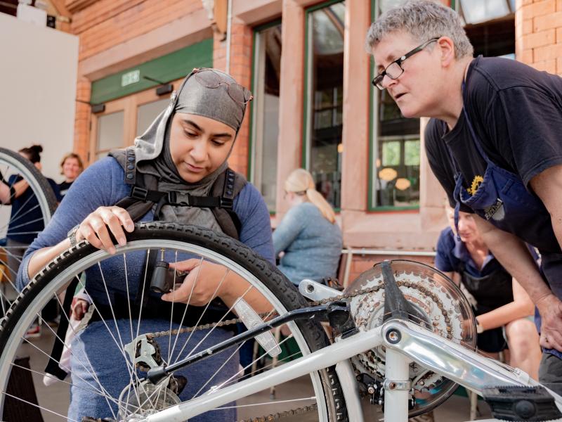A woman in a headscarf is fitting a bicycle pump to the valve on the rear tyre of an upside down bike. She is being watched by a man in overalls