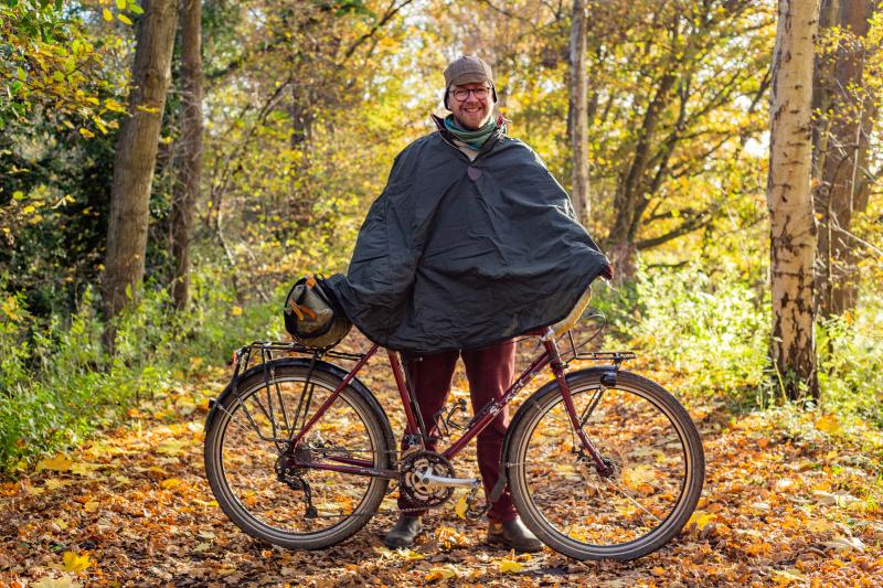 A man is standing on a woodland track holding a mountain bike. He is wearing a black rain poncho and smiling at the camera. It's autumn and the leaves are all turning gold and amber