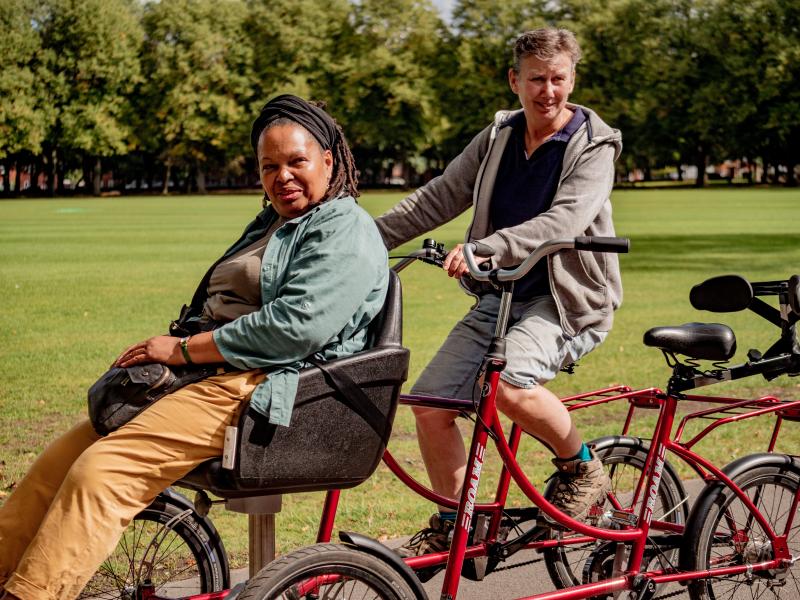 Two people are cycling on an adapted trike. A man is at the back pedalling, with a woman on a wheelchair adaptor at the front