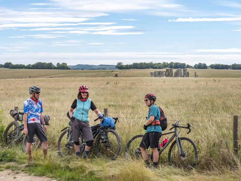 Three cyclists are standing at the edge of a field with their loaded touring bikes leaning against a wire fence. They are wearing cycling touring kit. They are all smiling. Stonehenge is in the background.