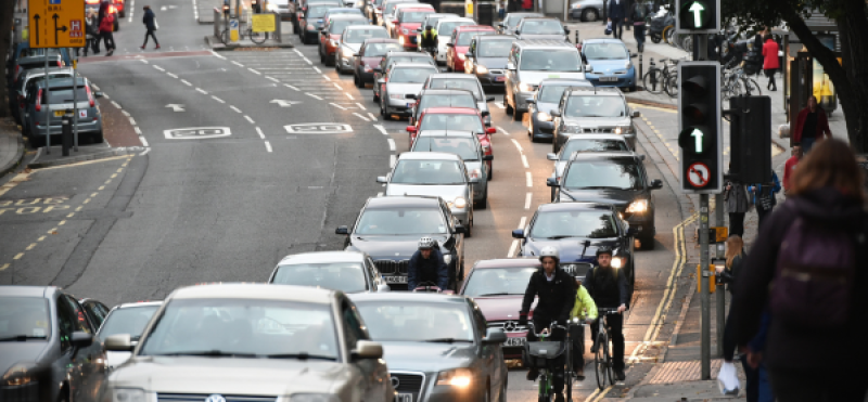 Two lines of stationary traffic are being passed by people on cycles on a busy urban road
