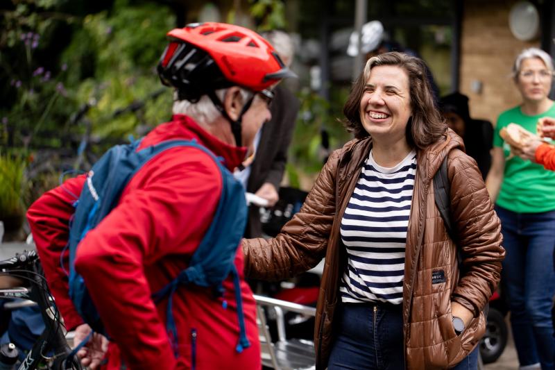 Two people are having a conversation. One is wearing a cycle helmet and jacket, the other is wearing normal clothes. They are laughing