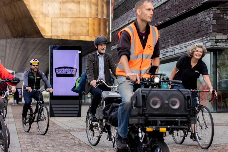 A group of people are riding on a shared-use path. A man in an orange hi-vis jacket is at the front