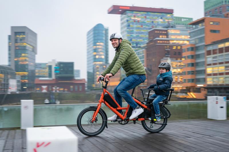 A man is riding an orange e-cargo bike with a child on the back, in an urban setting. He is wearing a green jacket and blue jeans, the child is wearing a blue jacket and jeans.