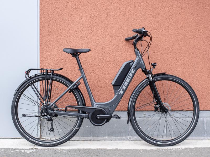 A grey hybrid e-bike with step-through frame, rear rack, flat handlebar and battery on the down tube, leaning against an orange and grey wall