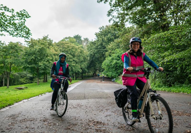 Two women are riding along a paved path in a park. They are wearing pink hi-vis vests