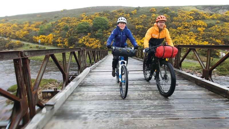 A man and a woman are cycling across a wooden bridge on loaded mountain bikes. It's very wet and they are wearing We are Cycling UK waterproofs