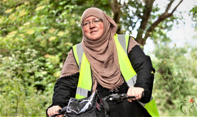 A woman in a hijab and head scarf with a high-vis jacket is cycling on a flat bar bike. She is smiling and wearing glasses