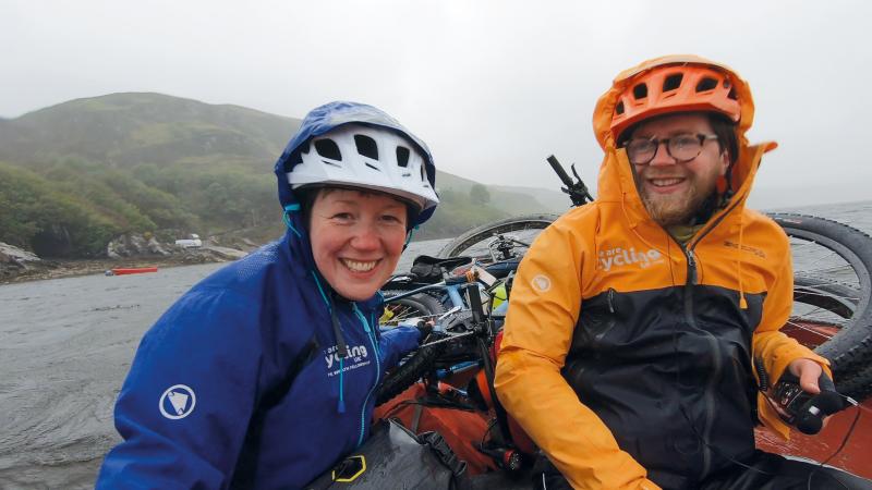 A man and a woman are on a ferry. They are both wearing We are Cycling UK waterproofs with hoods up over their cycling helmets and it's obviously raining. Their bikes can be seen behind them.
