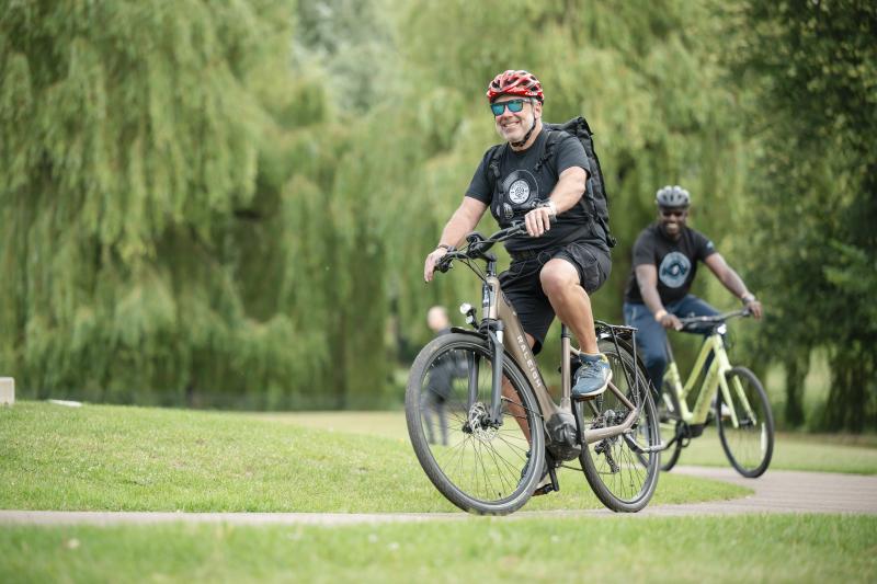 Two men are riding e-mountain bikes in a park. They are both wearing shorts and T-shirt and cycling helmets. It's summer and the trees are all very green. The two men are smiling
