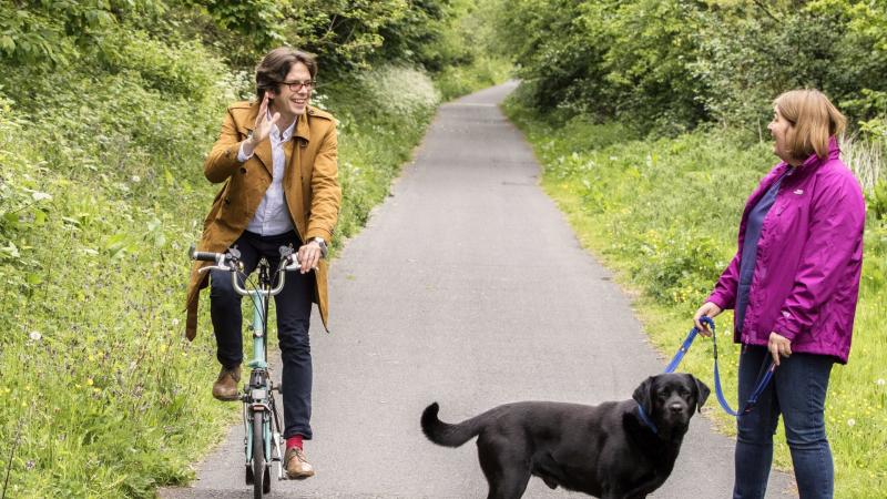 A man is cycling along a path on a Brompton, he is waving at a women with a black dog