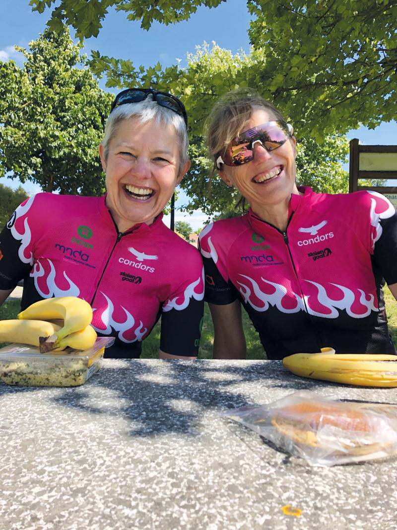 Two ladies in cycling jerseys sit on a bench in front of packed lunches
