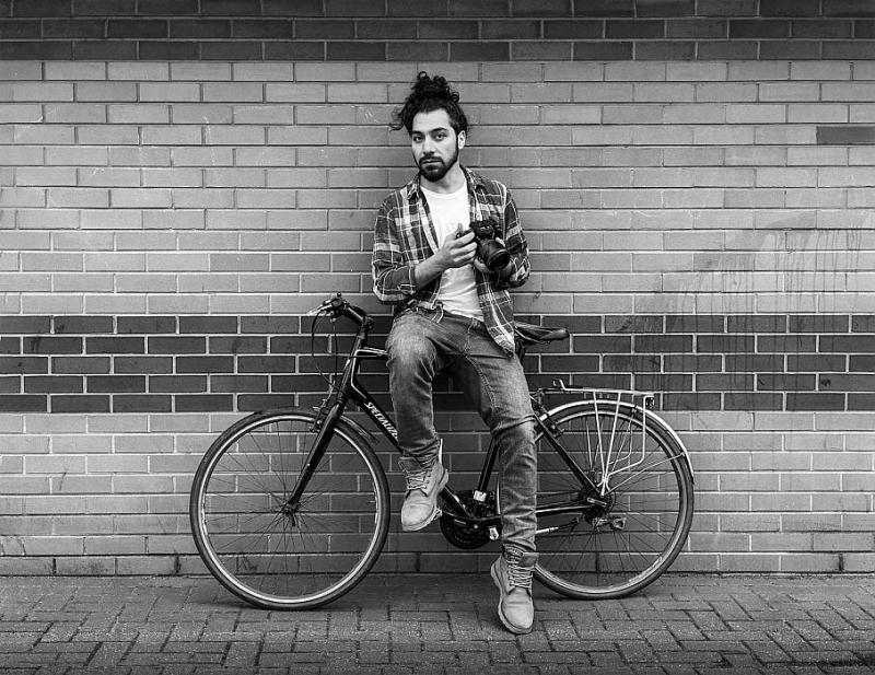 A man sits on a bicycle perched against a brick wall. He is wearing a check shirt over a white t-shirt and has his long hair tied up out of his face. He is holding a digital camera