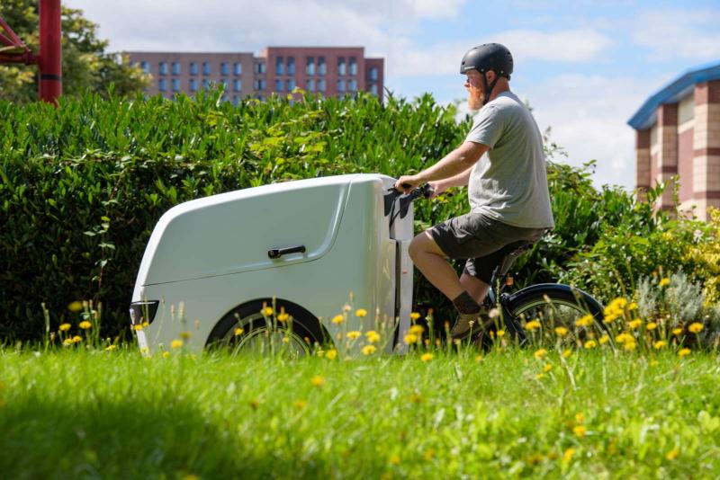 A man wearing a black helmet and grey t-shirt pedals a white e-cargo bike through grassy area on a sunny day