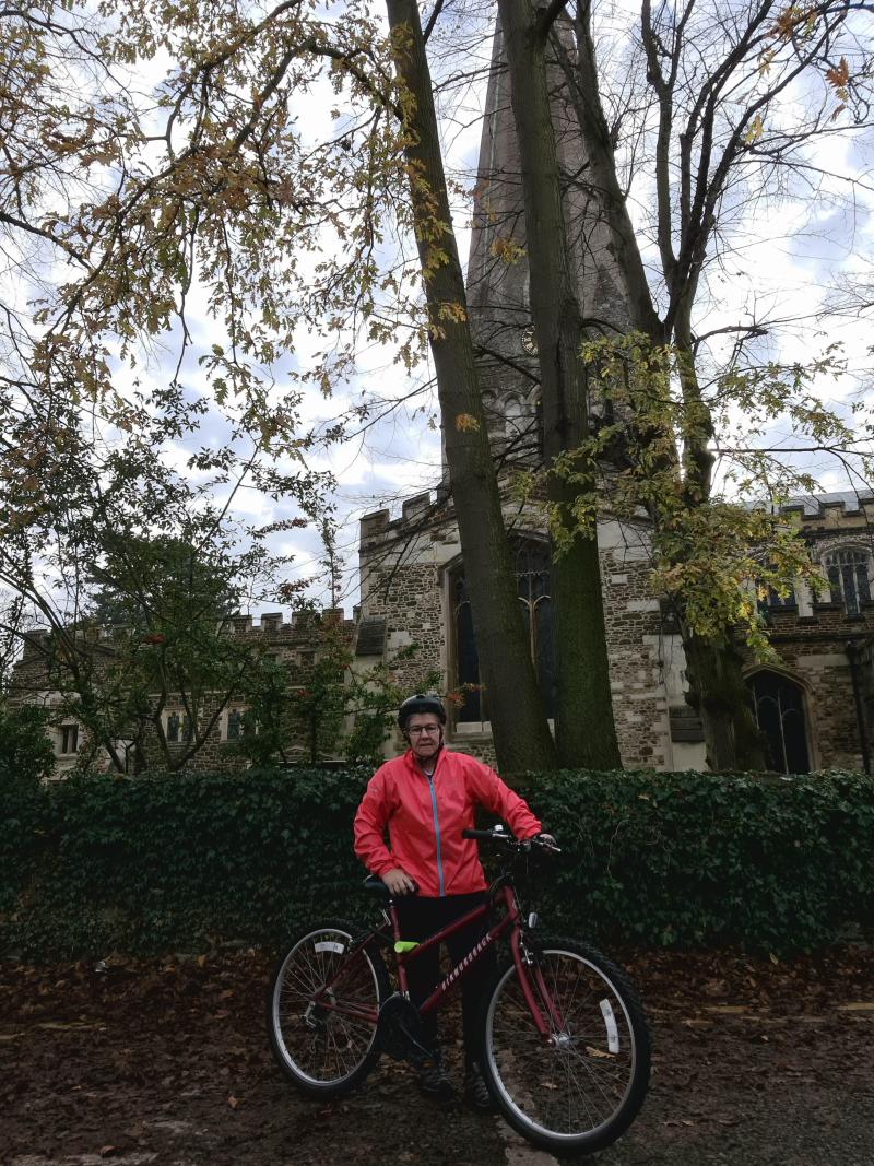 A middle-aged woman in red cycling kit stands by the roadside with a church tower in the background
